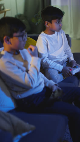 Vertikales-Video-Von-Zwei-Jungen,-Die-Zu-Hause-Auf-Dem-Sofa-Sitzen-Und-Spät-In-Der-Nacht-Mit-Einer-Computerspielkonsole-Im-Fernsehen-Spielen-Und-Controller-In-Der-Hand-Halten